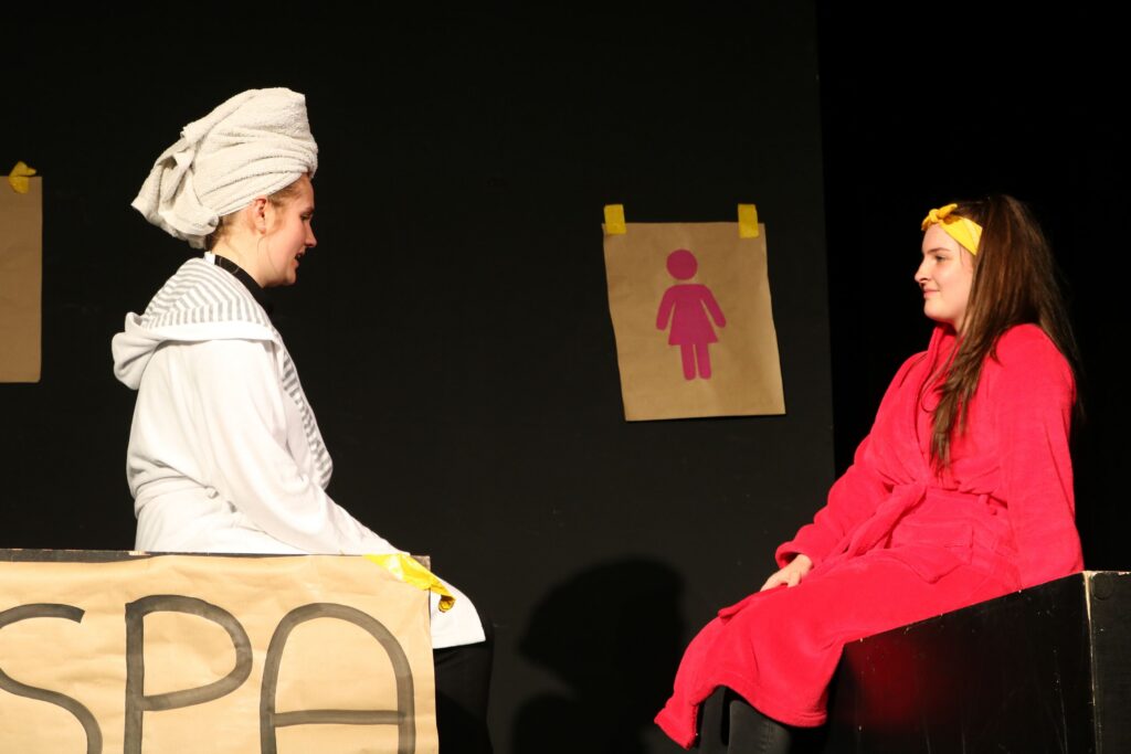 Zwei Schauspielerinnen in Bademänteln auf einer Bühne, hinter ihnen Spa-Dekoration.