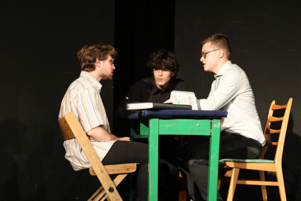 Drei Schüler sitzen an einem Tisch und diskutieren während einer Theateraufführung.
