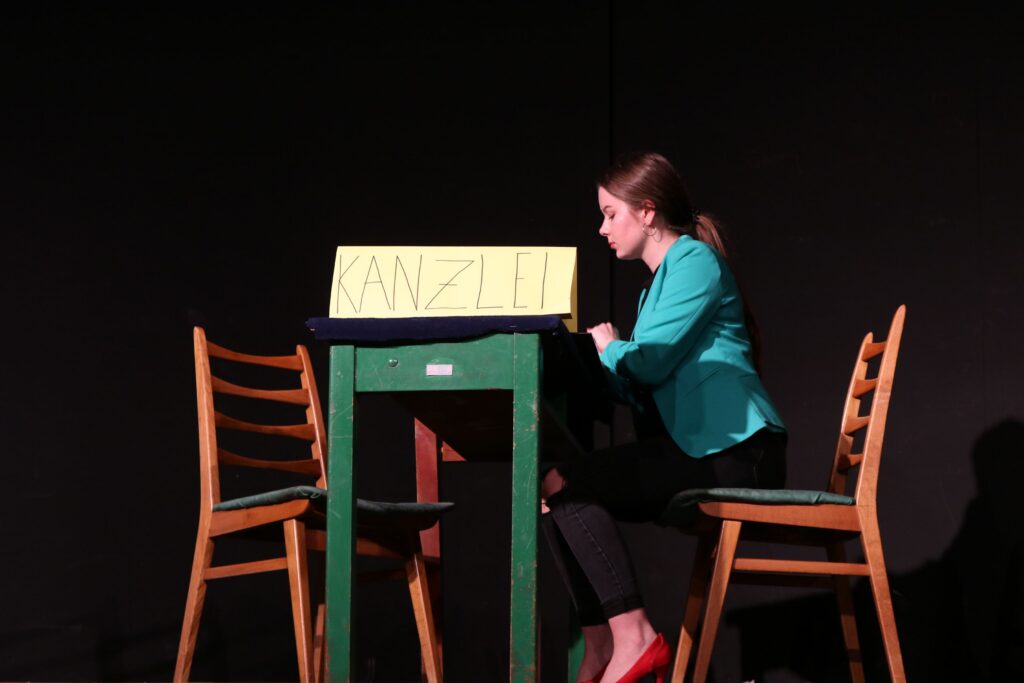 Eine Person sitzt an einem Tisch mit einem Schild "KANZLI". Zwei Stühle sind daneben.