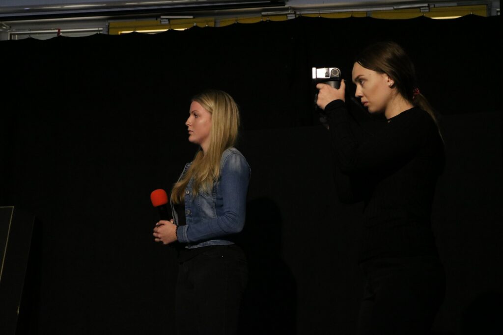 Zwei Mädchen auf einer Bühne; eines hält ein Mikrofon, das andere filmt.