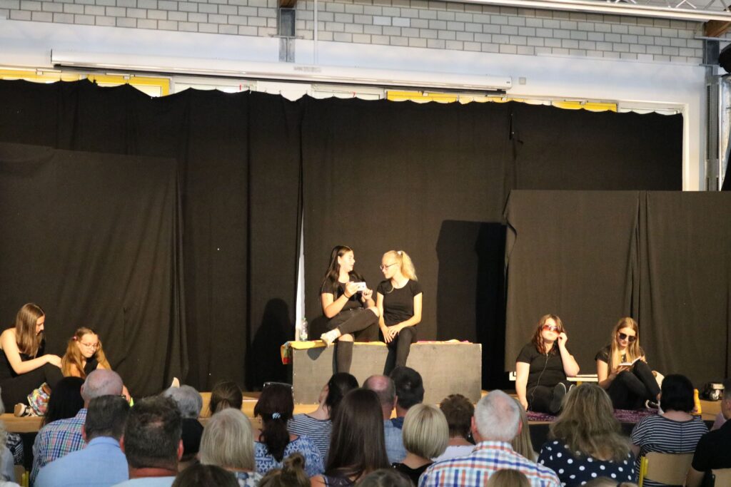 Theateraufführung mit Schauspielerinnen auf der Bühne vor Publikum.
