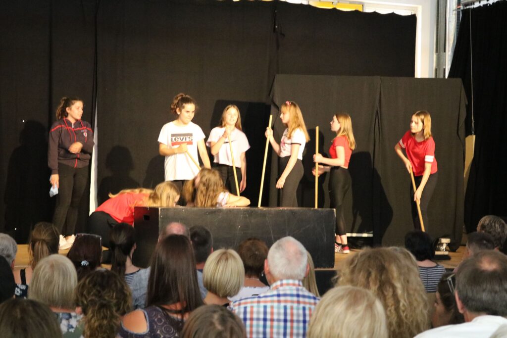 Schüler führen auf einer Bühne eine Theateraufführung auf. Zuschauer im Vordergrund.
