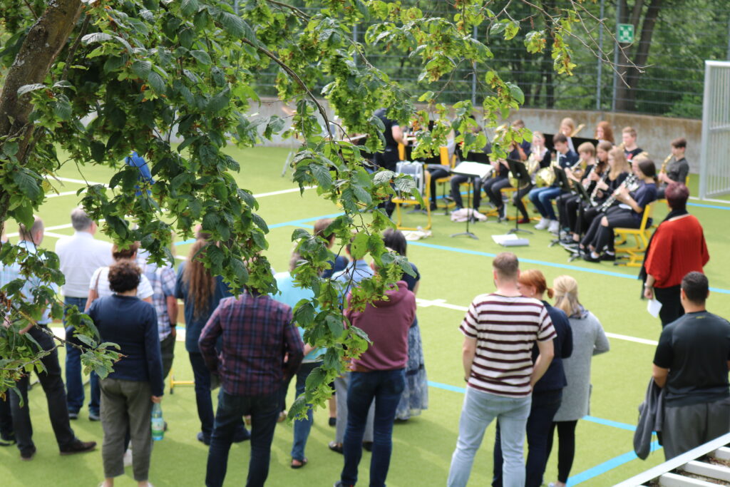 Besucher stehen vor einer Bandaufführung im Freien auf einem Sportplatz.