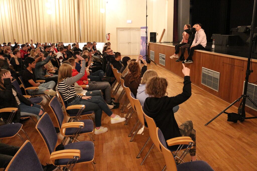 Schüler sitzen im Publikum und heben Hände während einer Aufführung.