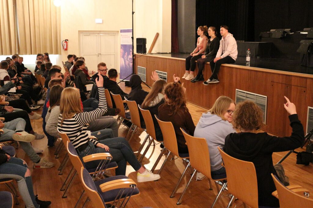 Schüler sitzen im Publikum, drei Personen auf der Bühne. Veranstaltungen in einem Saal.
