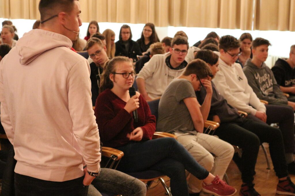 Schülerinnen und Schüler hören einem Vortrag zu, eine Schülerin stellt eine Frage.