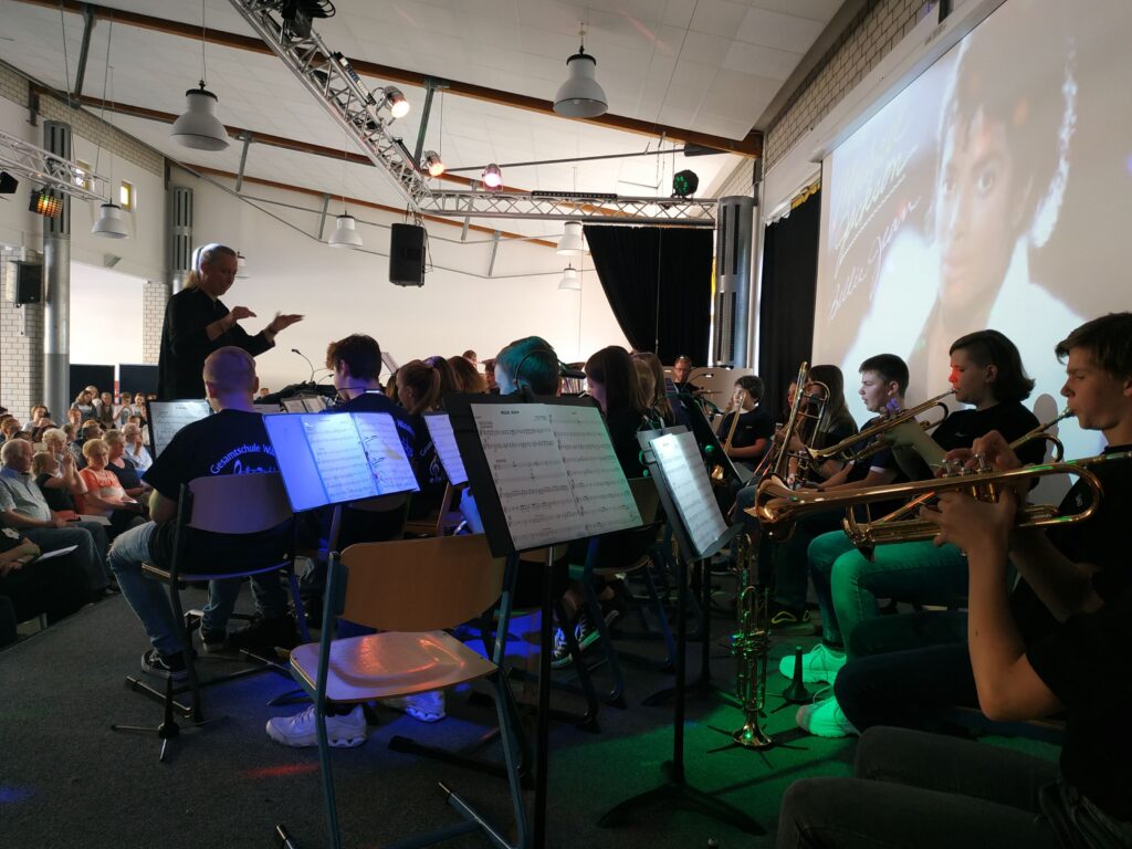 Konzert mit Orchester und Publikum, dirigiert von einer Person im Vordergrund.