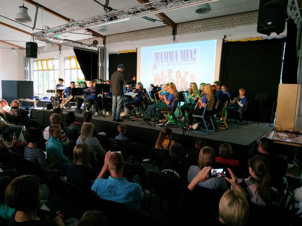 Schüler spielen in einer Aula ein Konzert, Publikum sitzt aufmerksam.