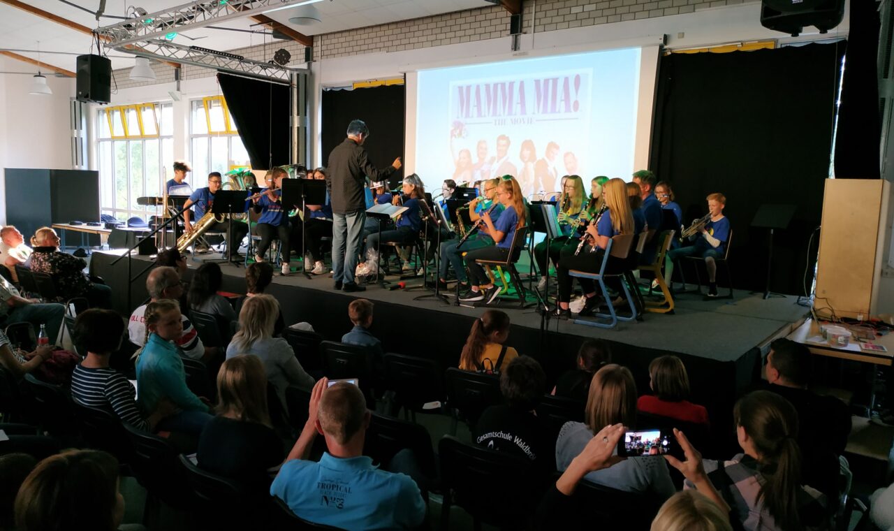 Schüler spielen in einer Aula ein Konzert, Publikum sitzt aufmerksam.