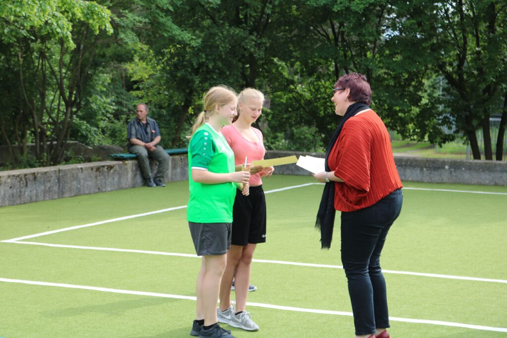 Zwei Mädchen erhalten eine Auszeichnung von einer Frau auf einem Tennisplatz.