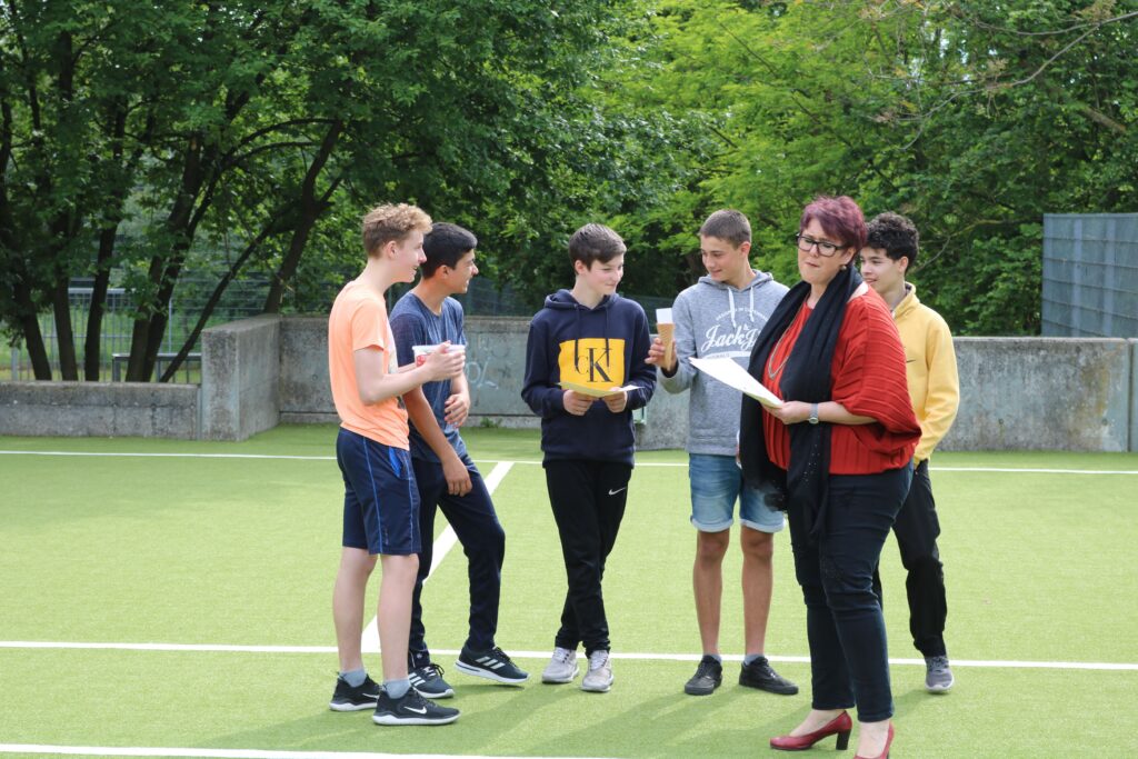 Schüler stehen auf dem Sportplatz und hören einer Lehrerin zu.