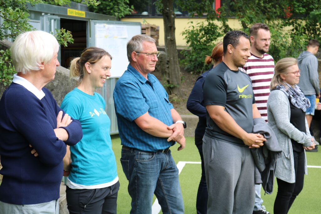 Gruppe von Erwachsenen steht auf einem Sportplatz und schaut aufmerksam zu.
