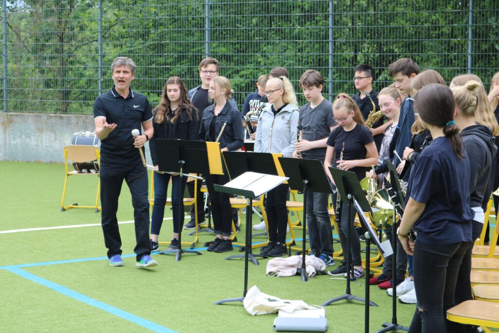 Ein Musikleiter dirigiert ein Ensemble auf einem Sportplatz.