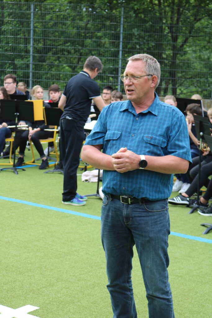 Ein Mann in blauer Hemd spricht vor einer Gruppe von Jugendlichen auf einem Sportplatz.