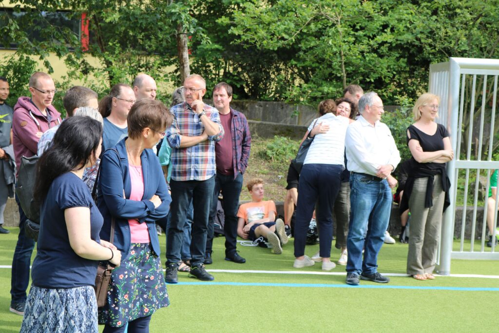 Gruppe von Menschen auf einem Schulgelände, einige stehen, andere sitzen im Hintergrund.