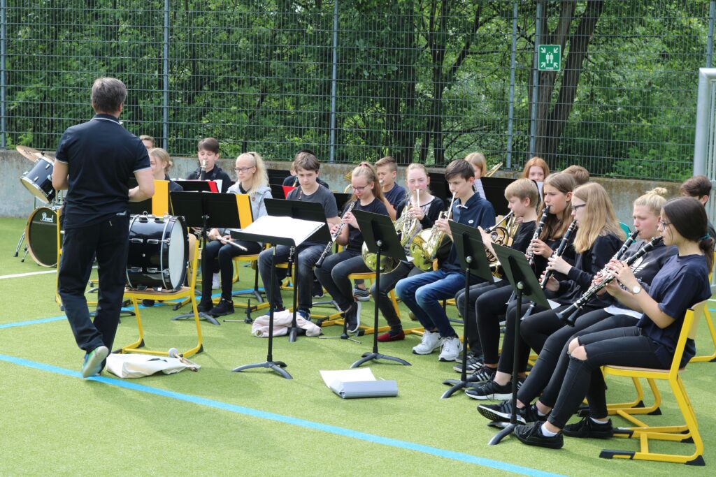 Schulblasmusikgruppe spielt auf einem Sportplatz, dirigiert von einem Lehrer.