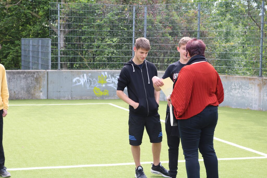 Schüler stehen auf einem Sportplatz und unterhalten sich mit einer Lehrkraft.