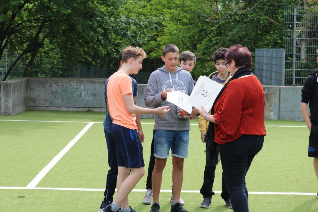 Schüler stehen in einer Gruppe auf dem Sportplatz und schauen auf ein Blatt Papier.