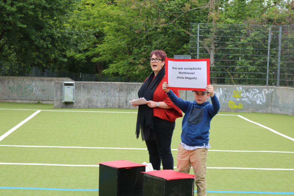 Frau mit Schild und Kind auf Sportplatz mit grüner Spielfläche.