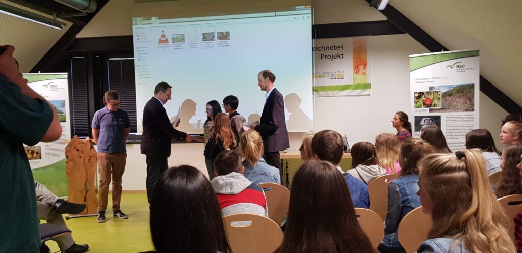 Zwei Männer übergeben einer Frau eine Auszeichnung auf einer Bühne vor Publikum.
