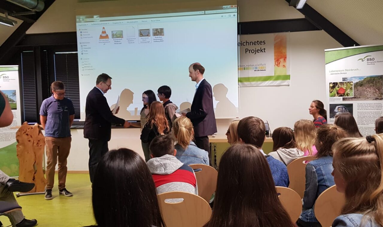 Zwei Männer übergeben einer Frau eine Auszeichnung auf einer Bühne vor Publikum.