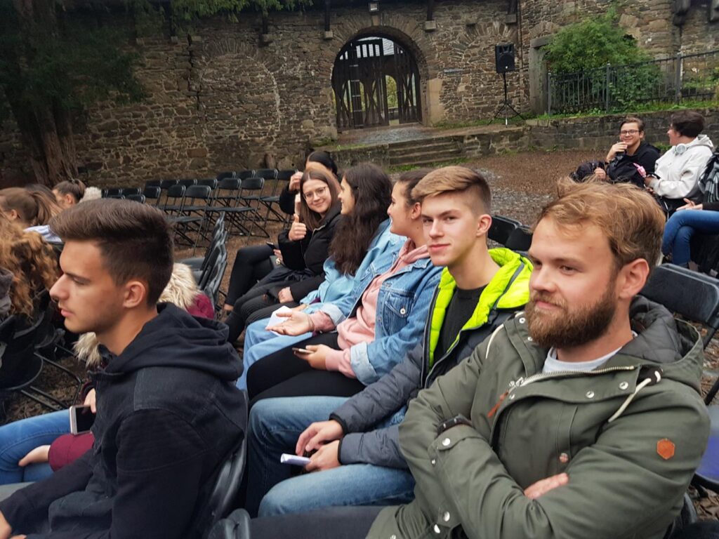 Gruppe von Jugendlichen sitzt auf Stühlen, ein älteres Gebäude im Hintergrund.
