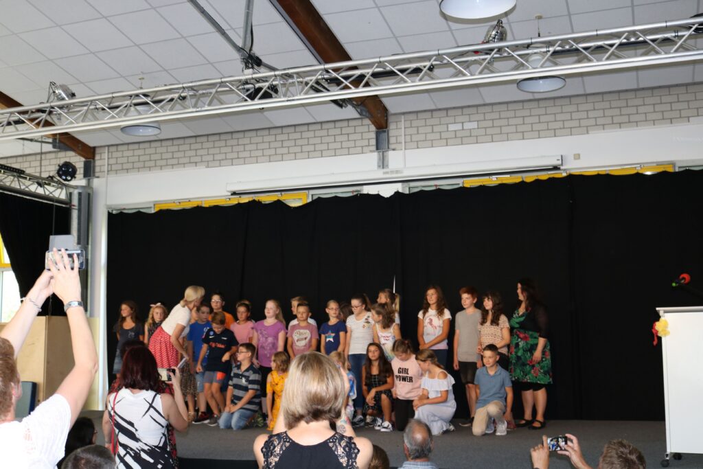 Schüler stehen auf einer Bühne bei einer Aufführung. Publikum im Hintergrund.