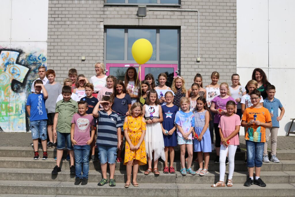 Schülergruppe mit Lehrern posiert vor Schulgebäude, gelbe Ballons im Hintergrund.