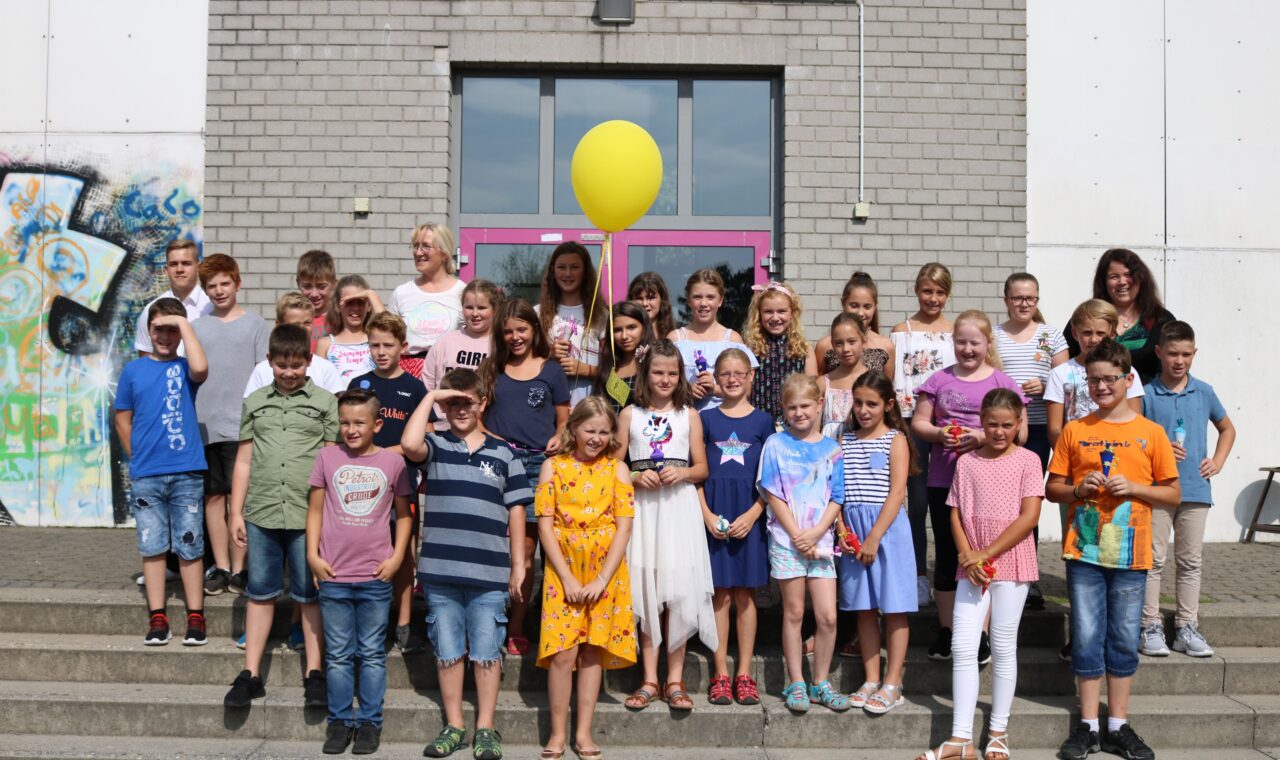 Schülergruppe mit Lehrern posiert vor Schulgebäude, gelbe Ballons im Hintergrund.