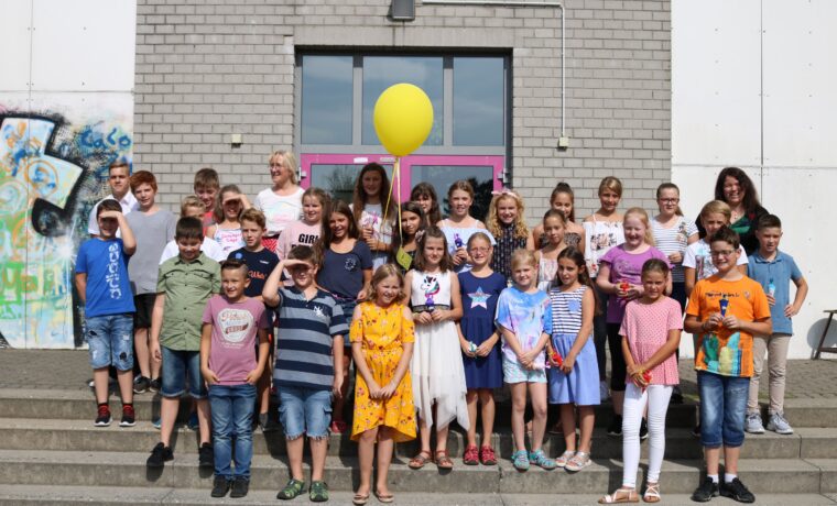 Schülergruppe mit Lehrern posiert vor Schulgebäude, gelbe Ballons im Hintergrund.