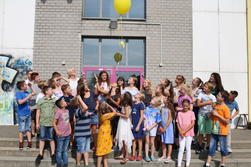 Gruppe von Schülern steht vor einem Gebäude und schaut auf gelbe Ballons.