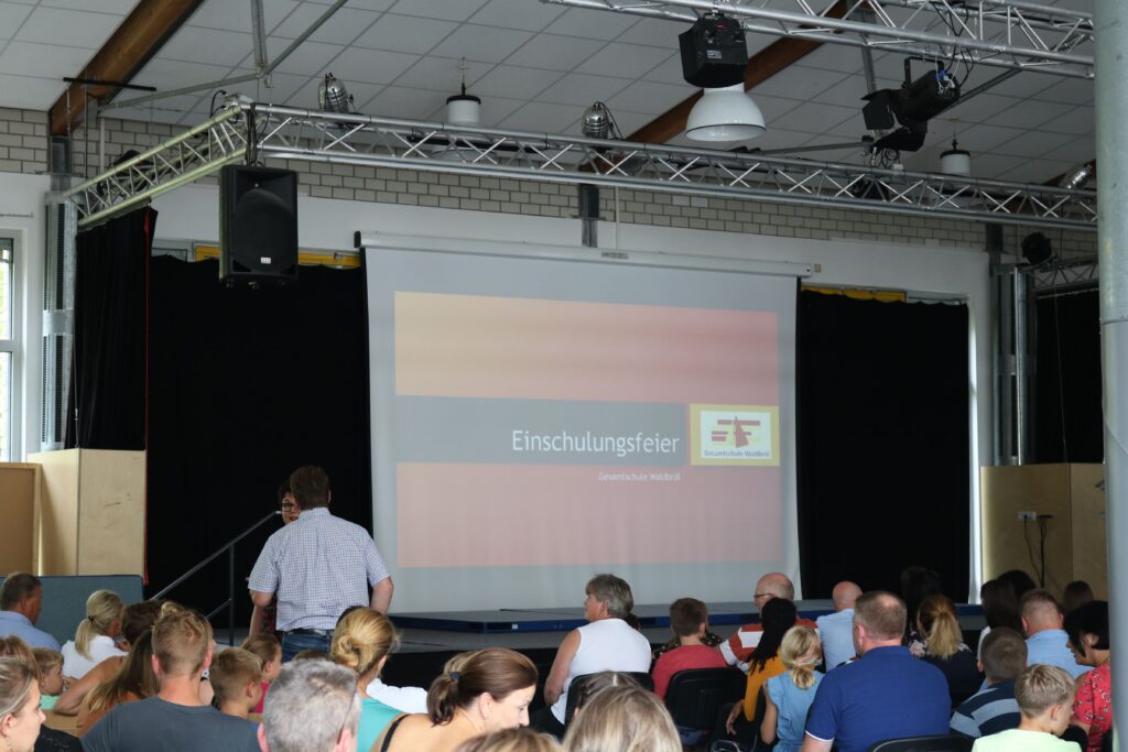 Bühne mit Leinwand und Publikum bei einer Veranstaltung in einem Saal.