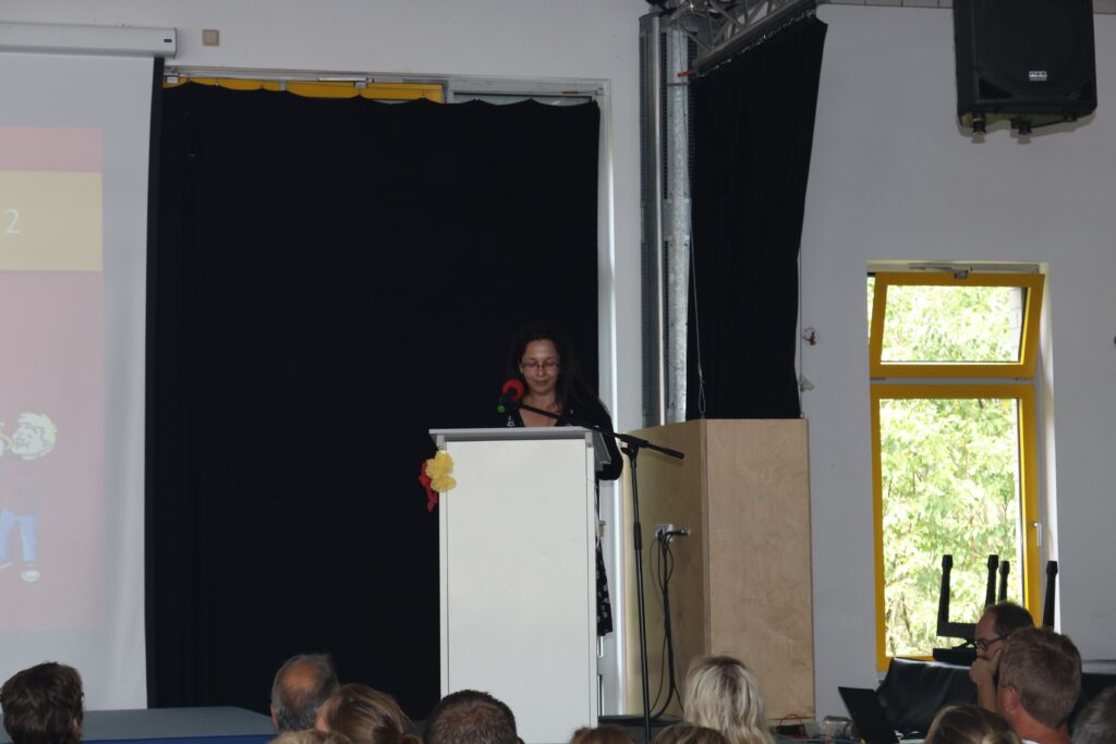 Person am Rednerpult vor einer Leinwand in einem Veranstaltungsraum.