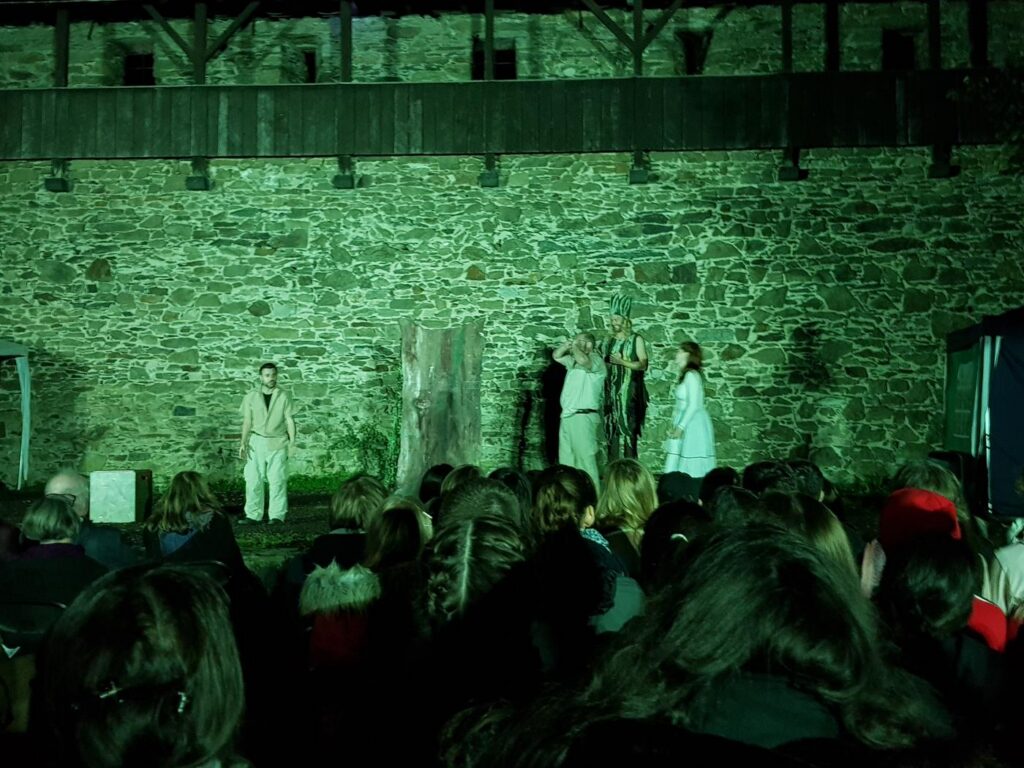 Theateraufführung vor einer historischen Steinmauer mit Zuschauern im Vordergrund.