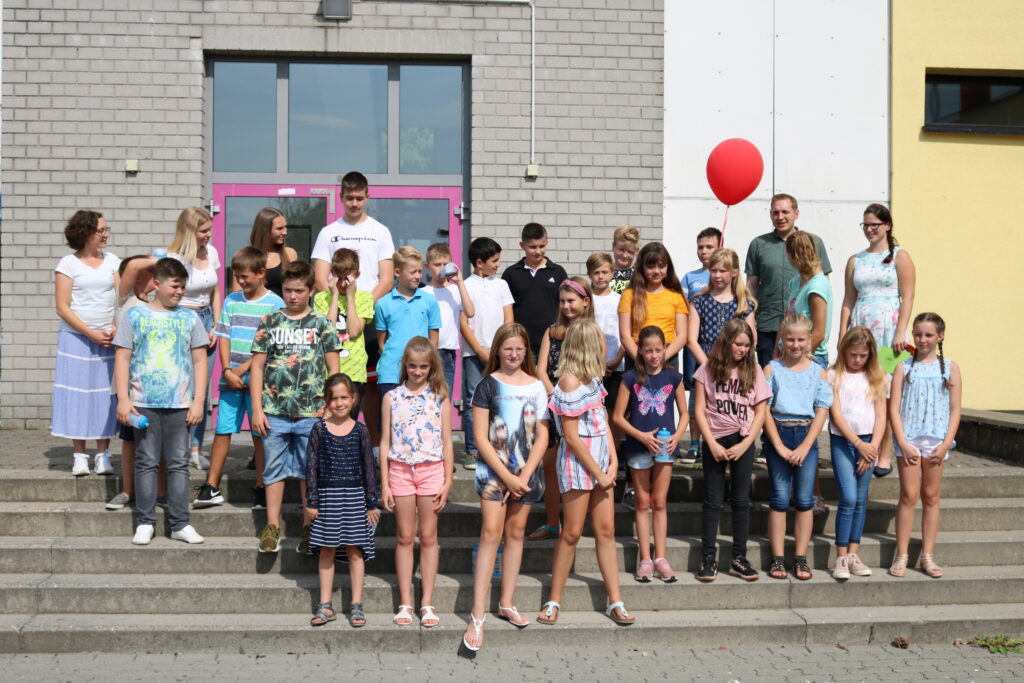 Gruppenfoto von Schülern und Lehrern vor der Schule, einige halten einen roten Luftballon.