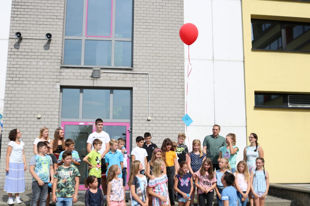 Schüler und Lehrer posieren vor einer Schule mit einem roten Luftballon.