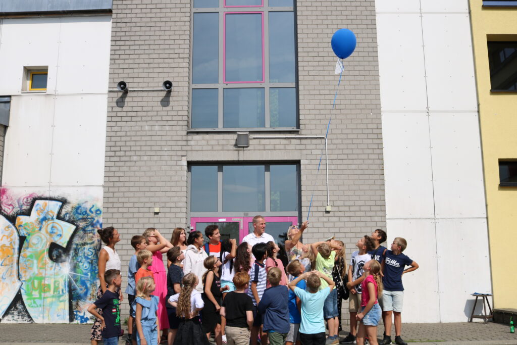 Eine Gruppe von Kindern steht vor einem Gebäude und schaut auf einen blauen Ballon.