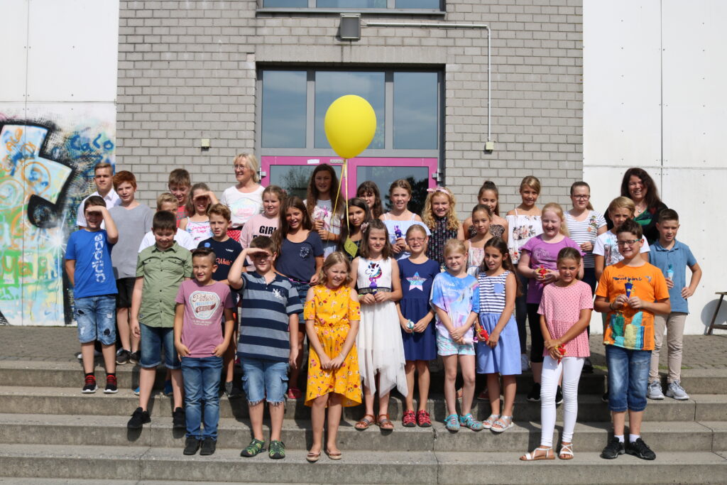 Schülergruppe steht auf Treppe vor einem Schulgebäude mit einem gelben Ballon.