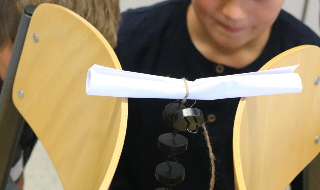 Schüler beobachten ein Experiment mit Stühlen und einem kleinen Objekt.