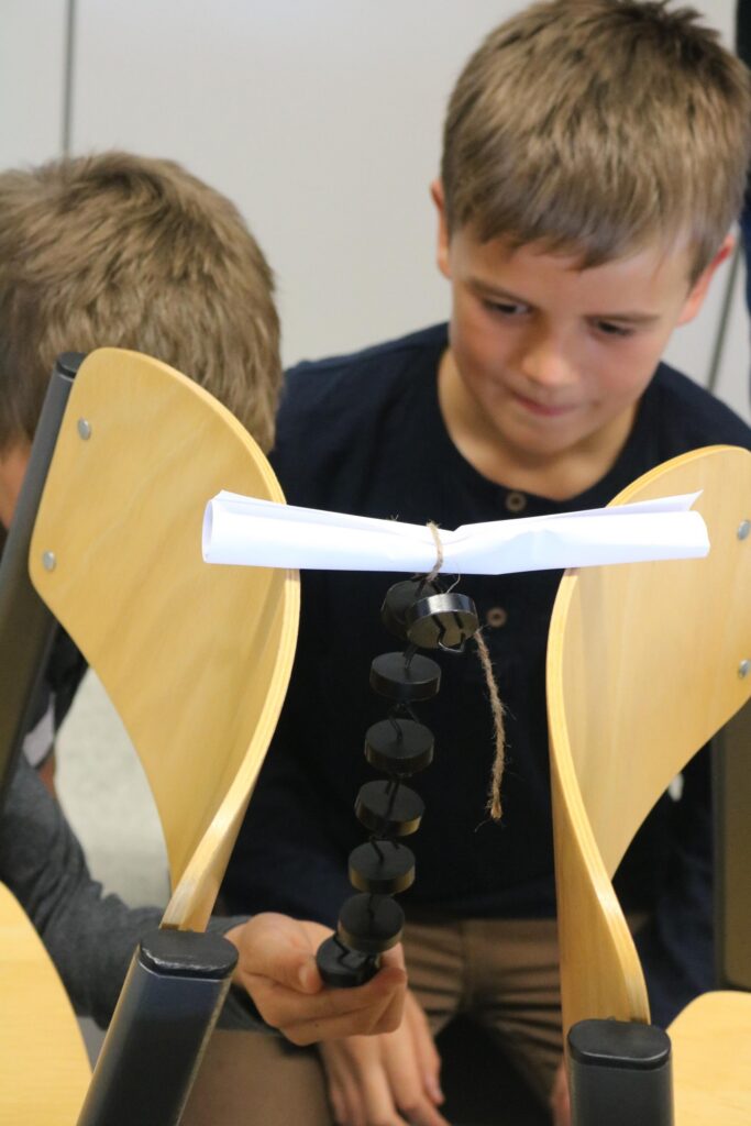 Schüler beobachten ein Experiment mit Stühlen und einem kleinen Objekt.