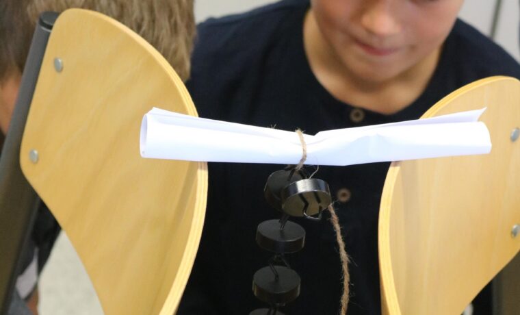 Schüler beobachten ein Experiment mit Stühlen und einem kleinen Objekt.
