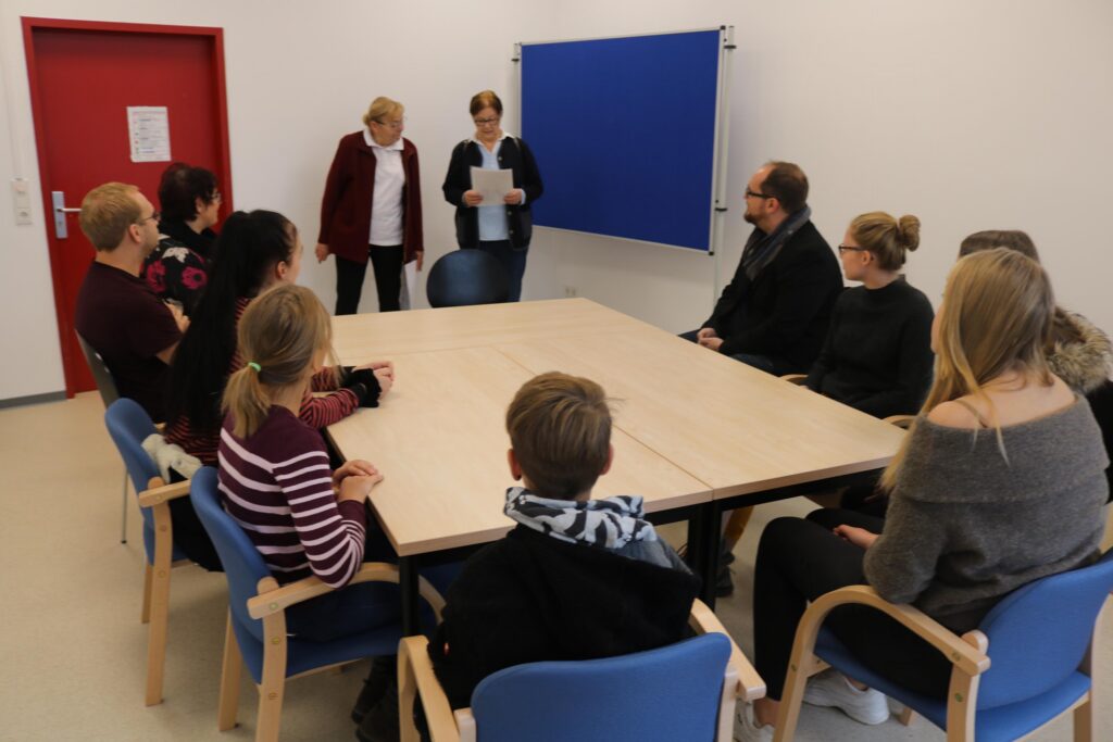 Personen sitzen um einen Tisch in einem Besprechungsraum. Zwei Personen stehen.