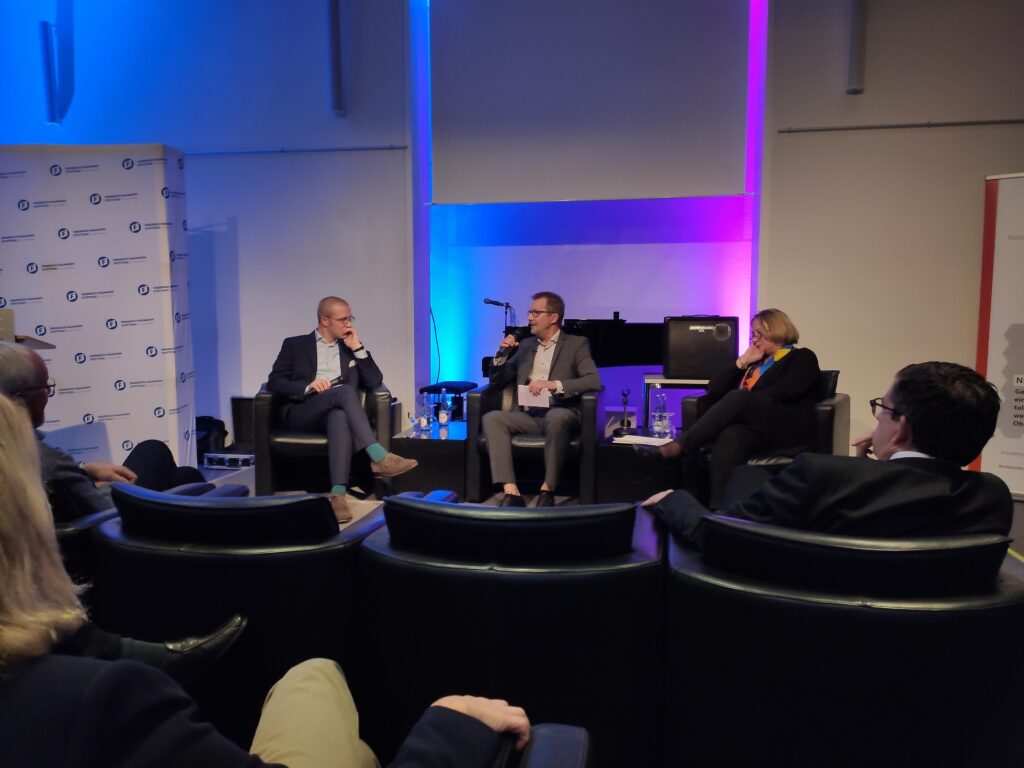 Podiumsdiskussion mit drei Personen auf einer Bühne in einem Seminarraum.