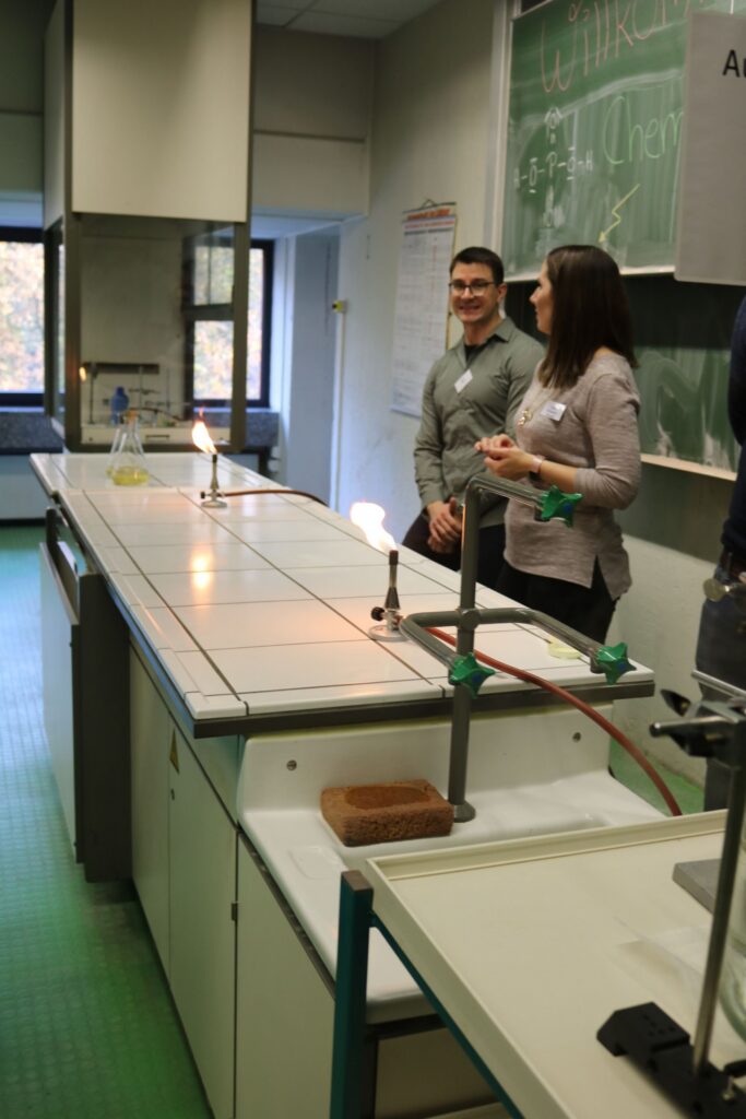 Zwei Personen stehen an einem Tisch in einem modernen Unterrichtsraum mit Experimentierstationen.
