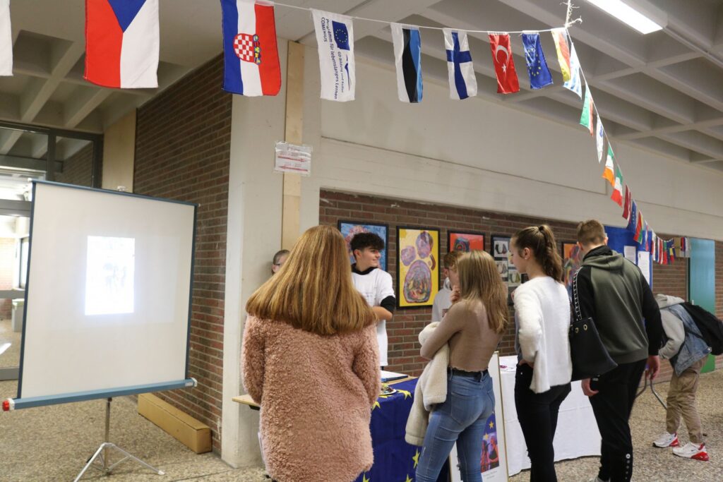 Schüler stehen an einem Informationsstand mit Fahnen und Plakaten im Schulgebäude.