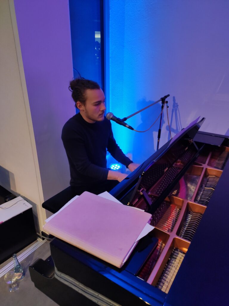 Ein junger Mann spielt Klavier und singt in einem blau beleuchteten Raum.