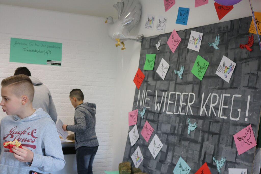 Schüler stehen vor einer Wand mit bunten Papierbotschaften und dem Schriftzug "Nie wieder Krieg!".