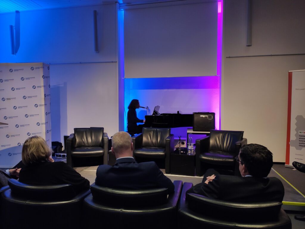 Eine Pianistin spielt an einem Klavier vor einer Zuschauergruppe auf schwarzen Sesseln.