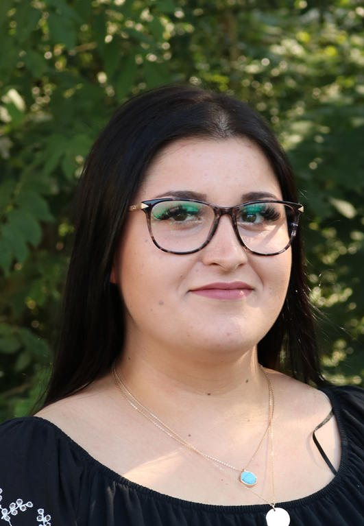 Frau mit Brille steht vor einer grünen, natürlichen Kulisse.