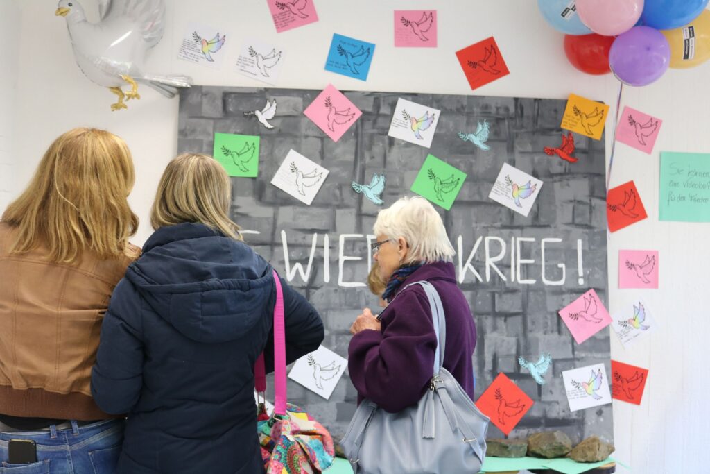 Drei Personen betrachten ein Wandbild mit bunten Notizen und dem Schriftzug „WIE IM KRIEG!“.